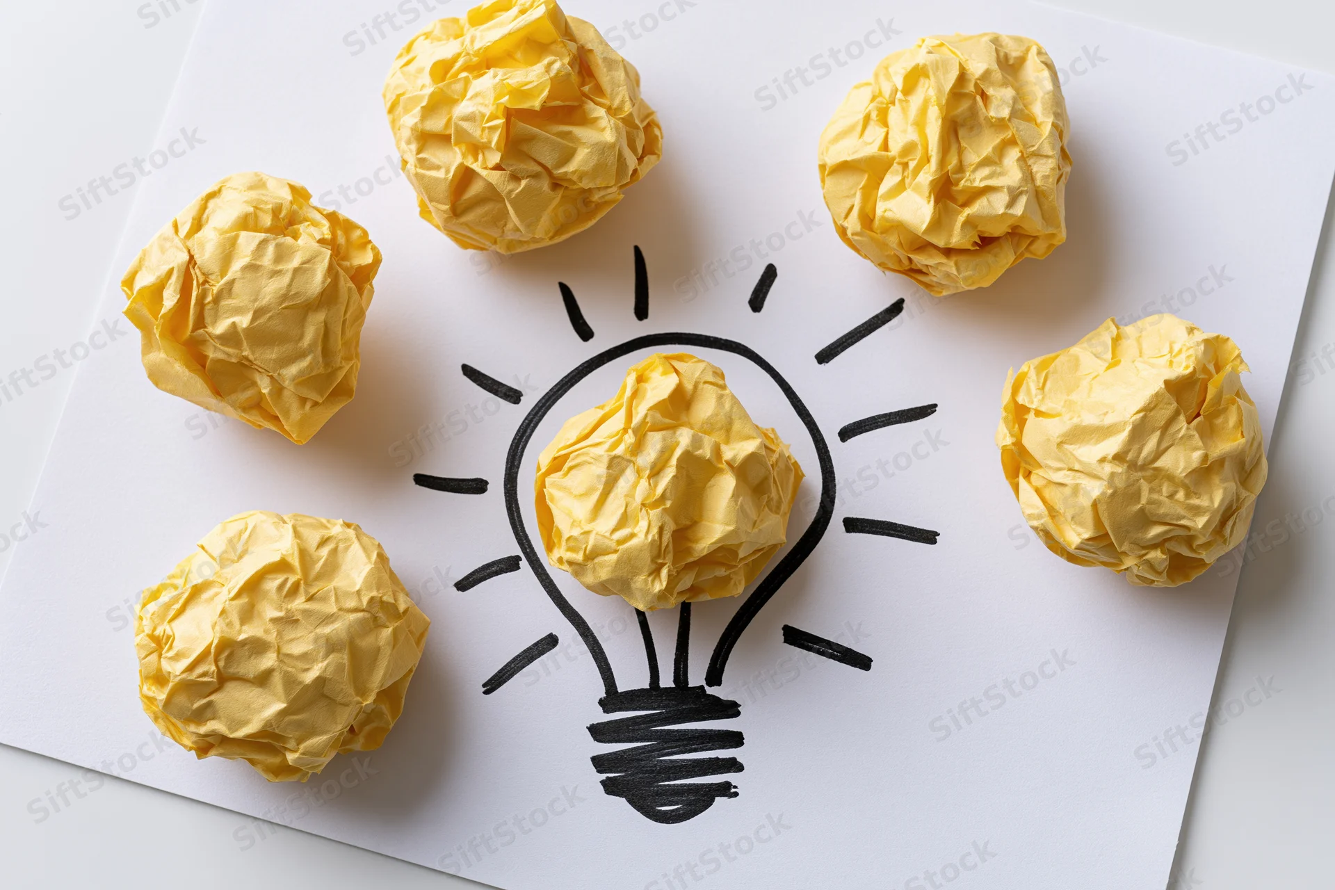 Crumpled yellow paper balls arranged around a hand-drawn lightbulb on white paper