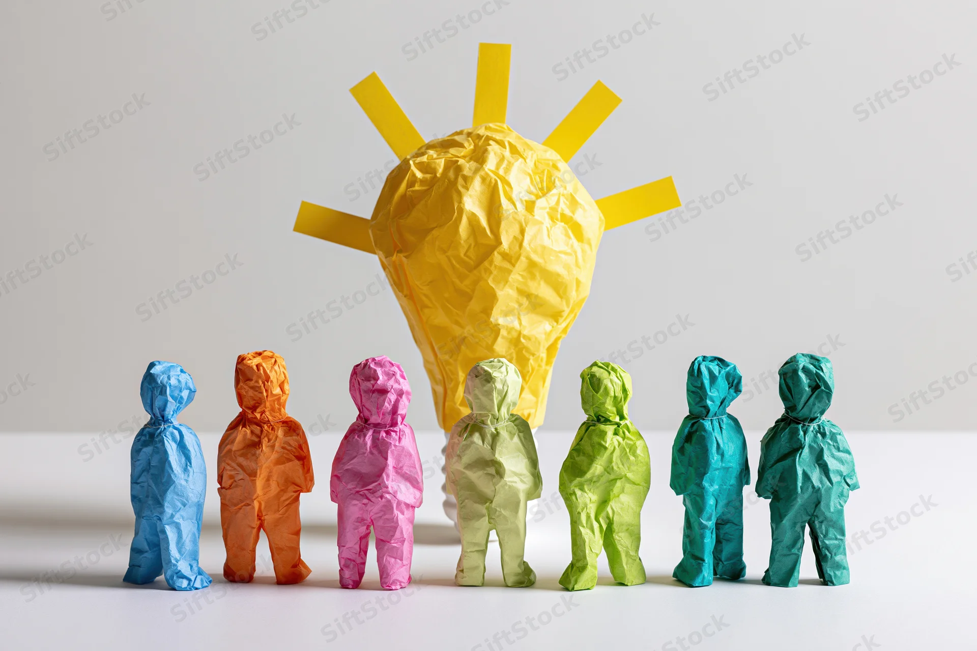 Colorful crumpled paper figures standing in a row facing a large yellow paper lightbulb on a white surface