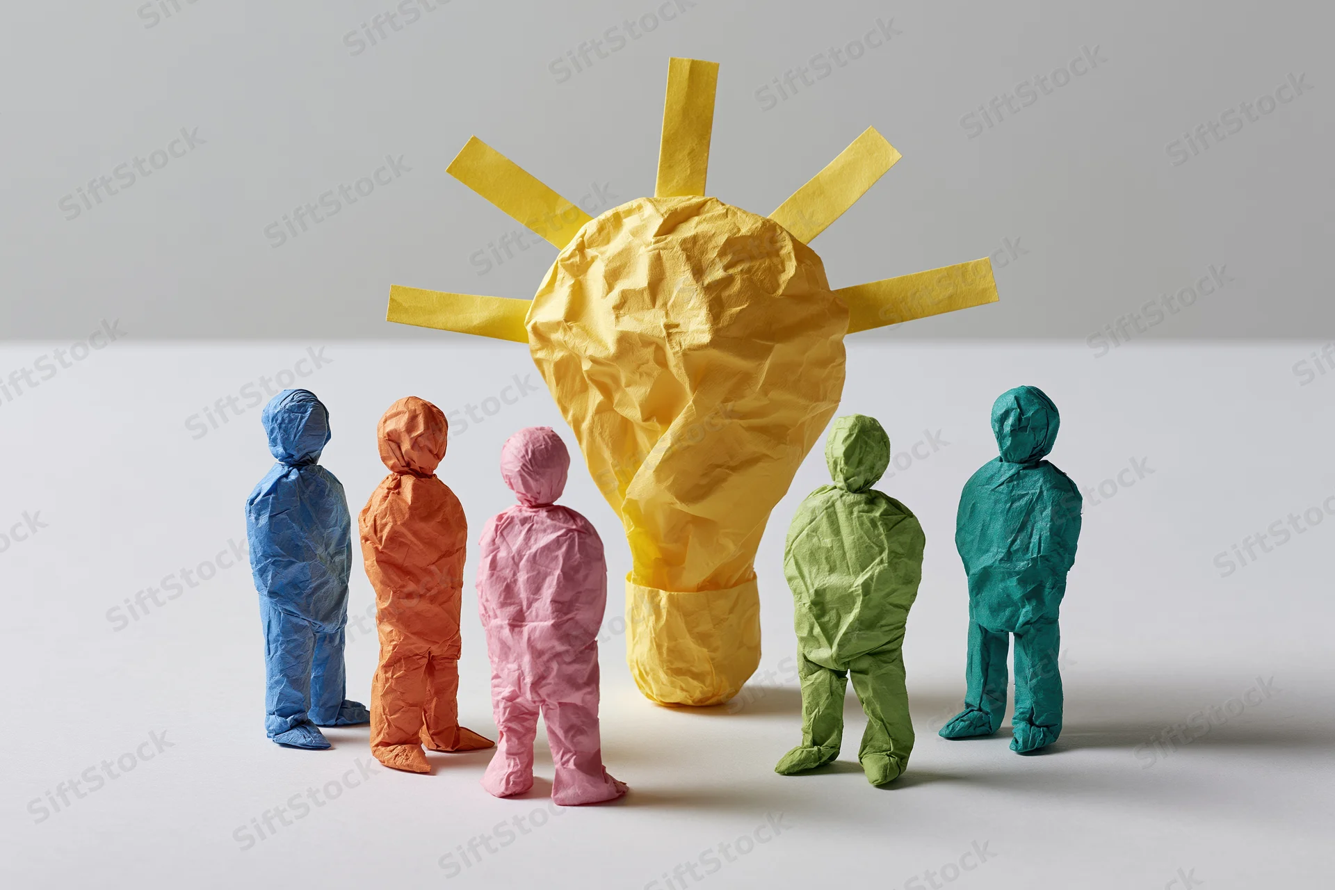 Five colorful paper figures standing around a large crumpled yellow lightbulb on a white surface