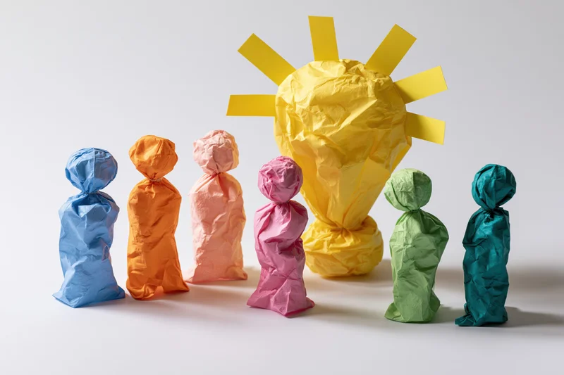 Crumpled paper figures in bright colors arranged around a yellow lightbulb sun symbol on white surface