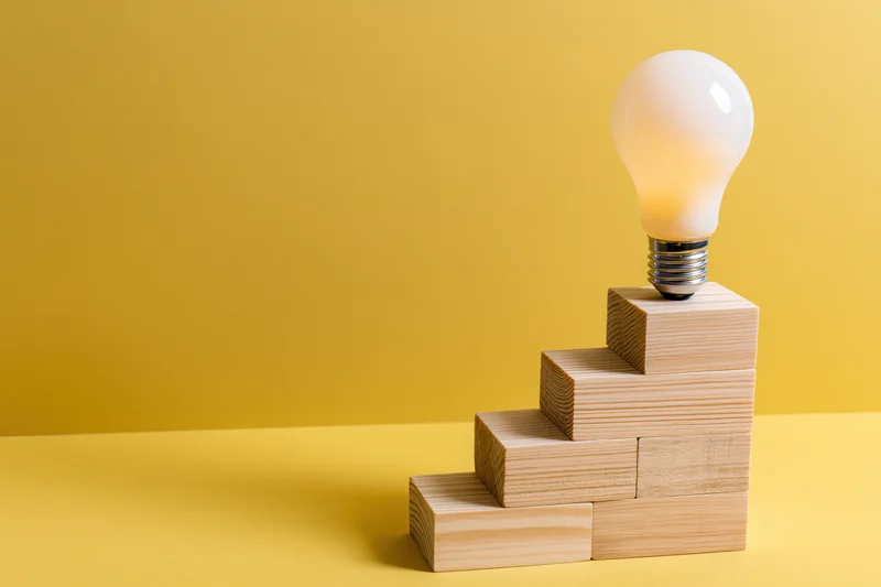 Lightbulb resting on top of wooden blocks arranged as ascending staircase against yellow background