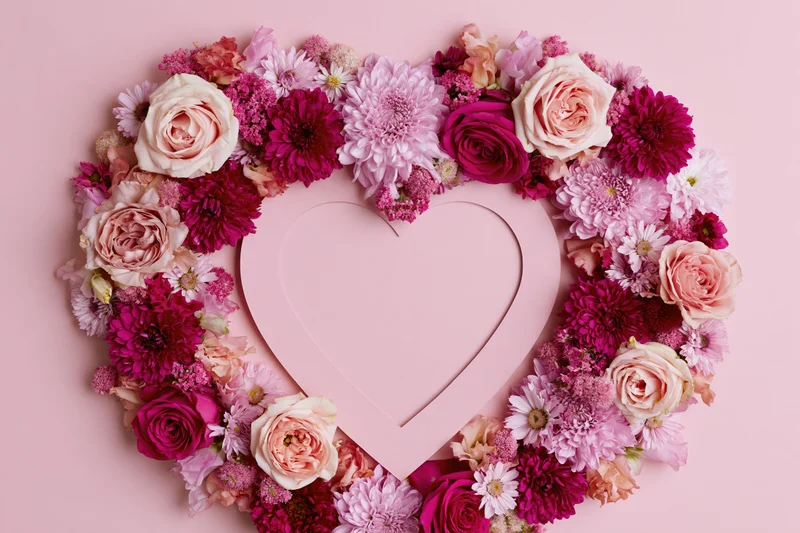 Heart-shaped floral arrangement made of pink and magenta roses and dahlia flowers on a pink background
