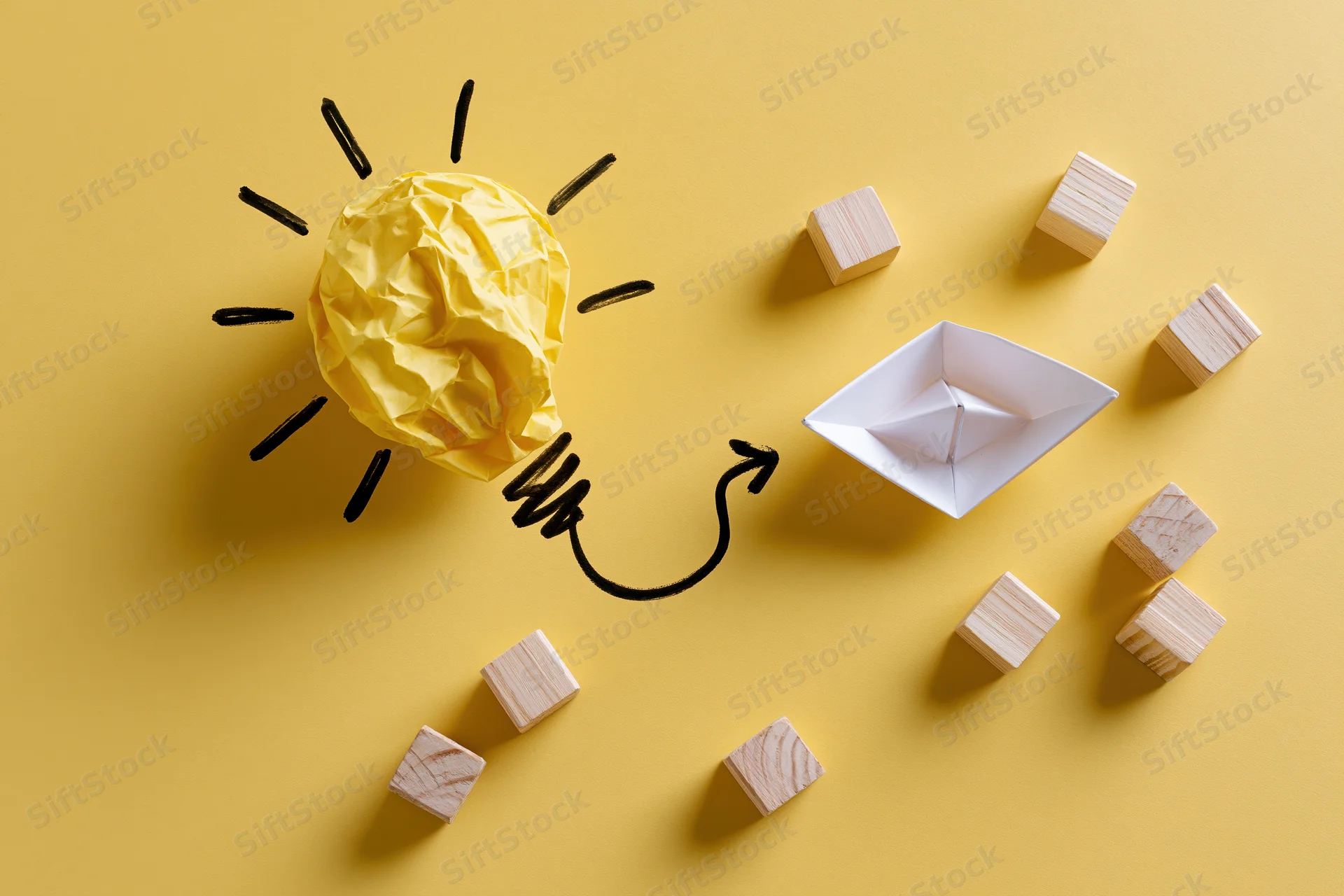 Crumpled yellow paper shaped as a lightbulb with hand-drawn rays and an arrow pointing to a paper boat surrounded by wooden cubes on a yellow surface