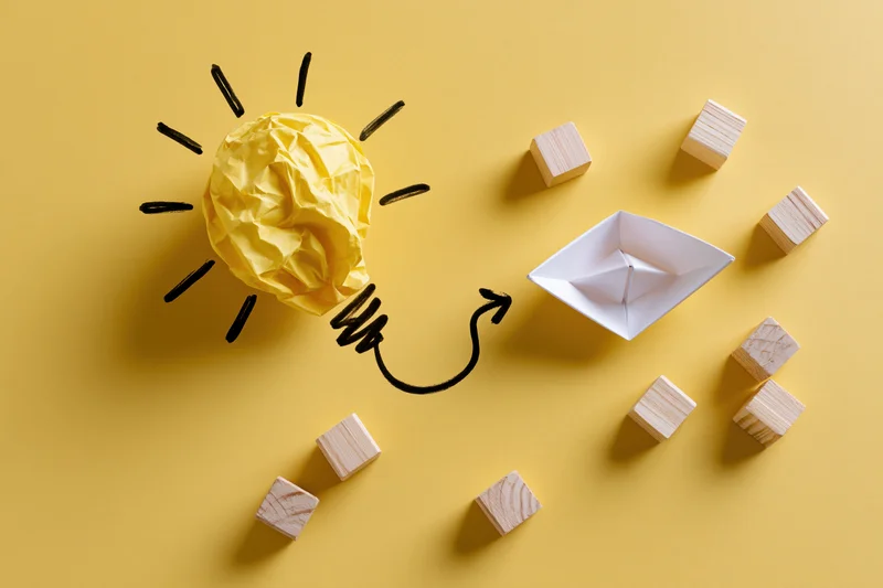 Crumpled yellow paper shaped as a lightbulb with hand-drawn rays and an arrow pointing to a paper boat surrounded by wooden cubes on a yellow surface