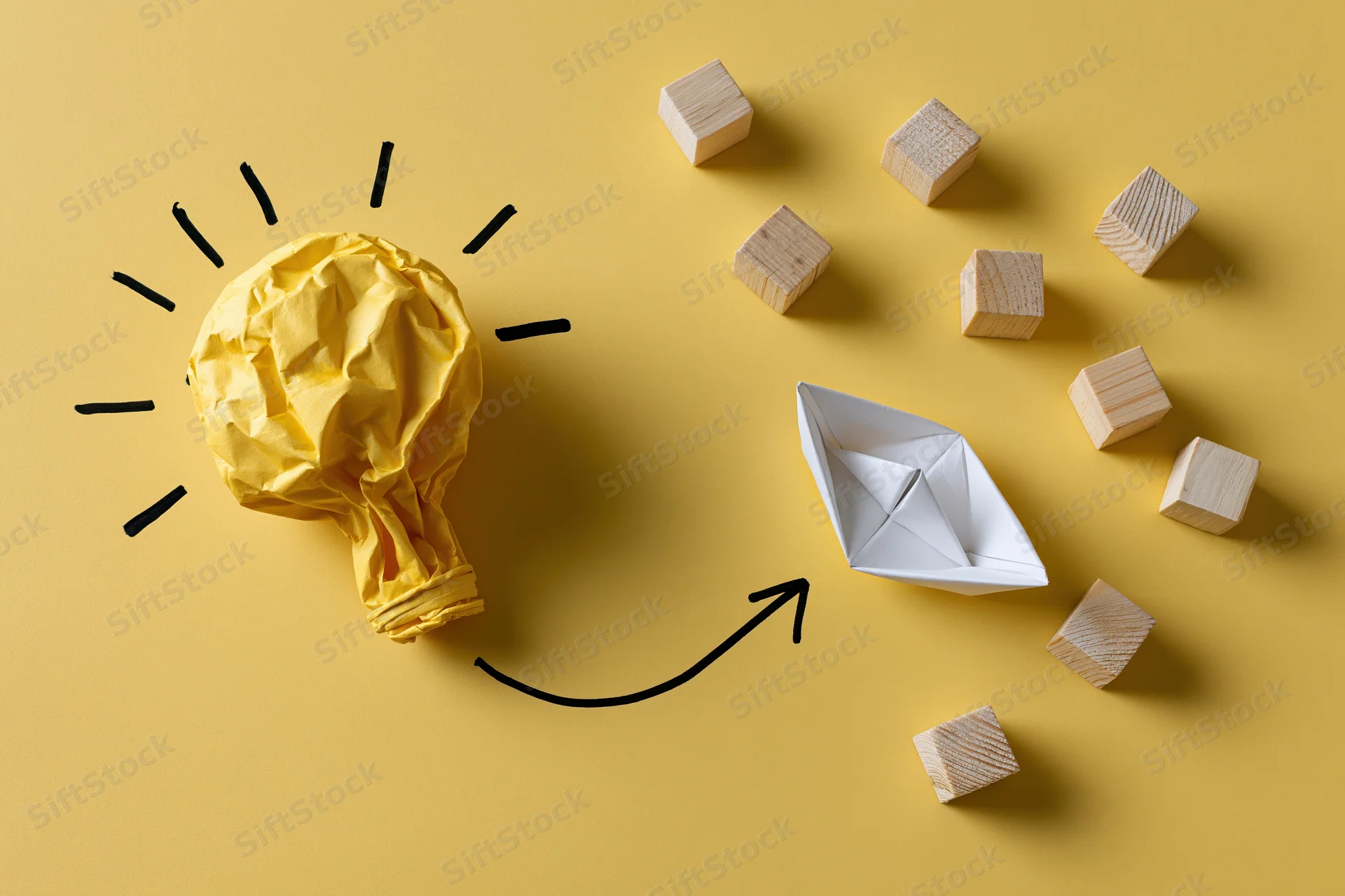Crumpled yellow paper lightbulb with hand-drawn rays and arrow pointing to white origami boat surrounded by wooden cubes on yellow background