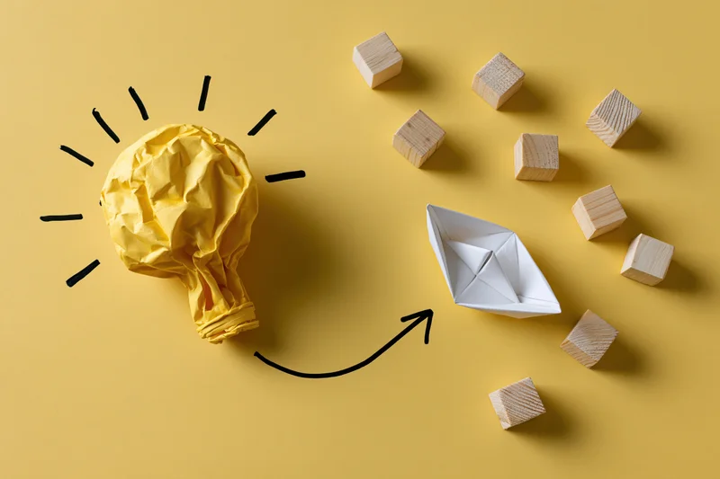 Crumpled yellow paper lightbulb with hand-drawn rays and arrow pointing to white origami boat surrounded by wooden cubes on yellow background