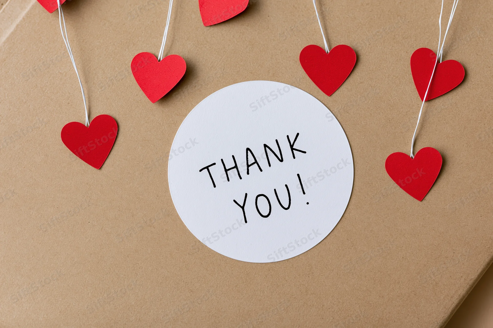 Round white sticker with thank you text on brown kraft paper surrounded by red hearts on strings