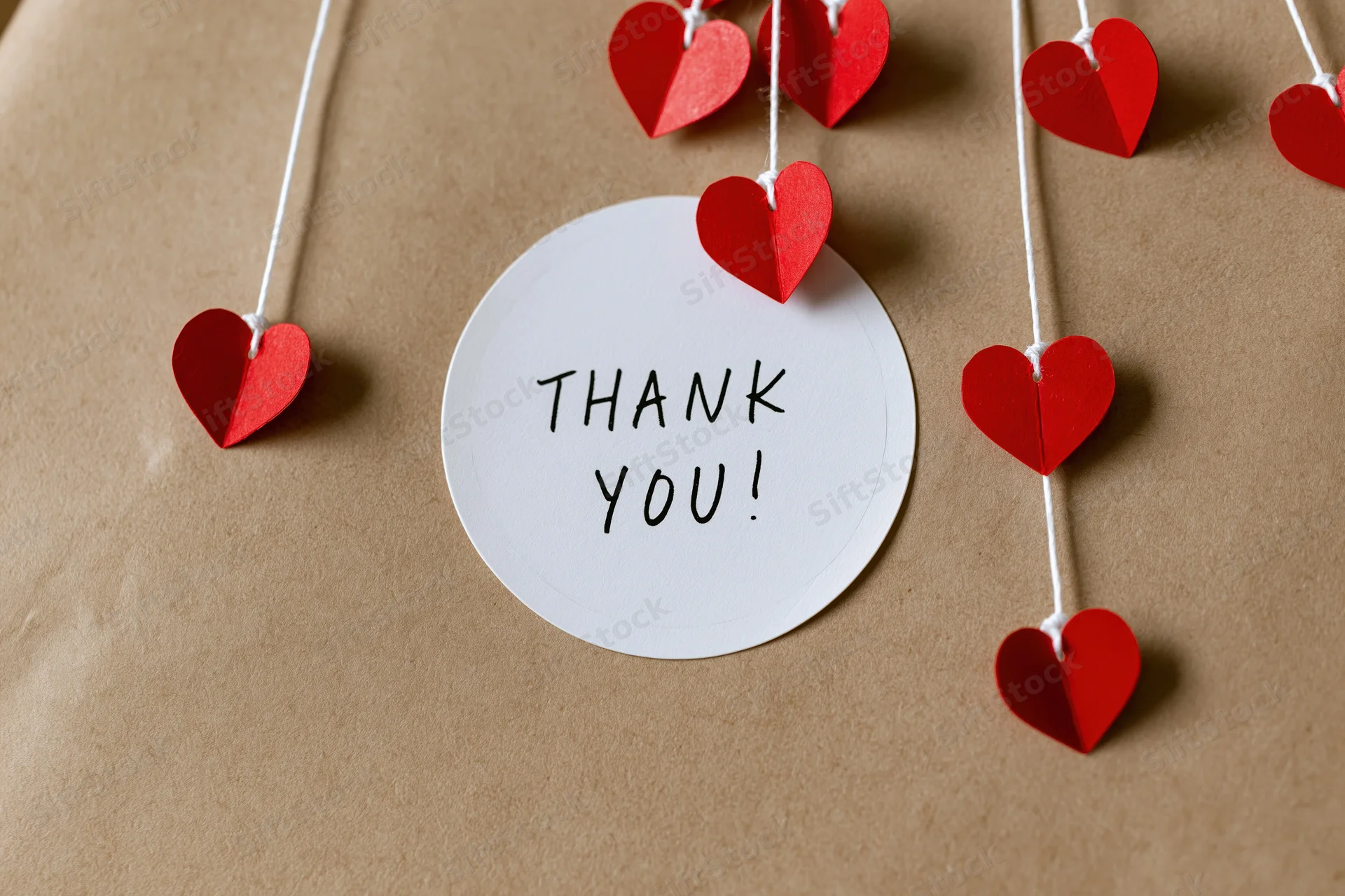 Round white sticker with thank you text surrounded by red paper hearts on string over brown kraft paper