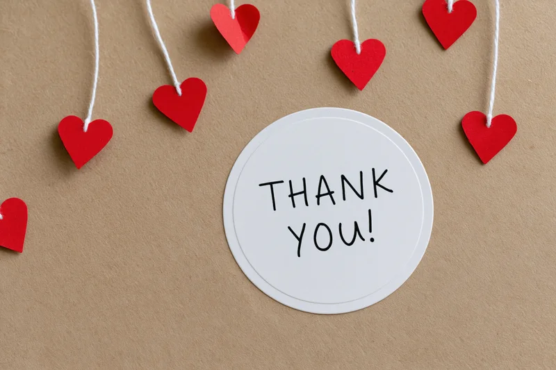 Round white sticker with thank you text surrounded by red hearts on string on brown paper surface