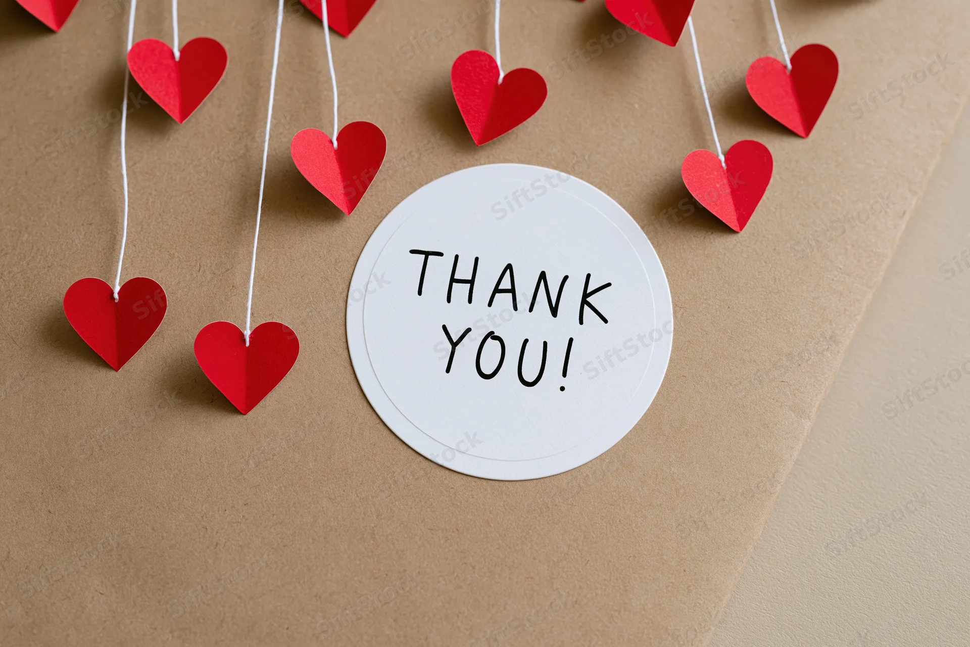 Round white sticker with thank you text on brown paper surrounded by red paper hearts on strings