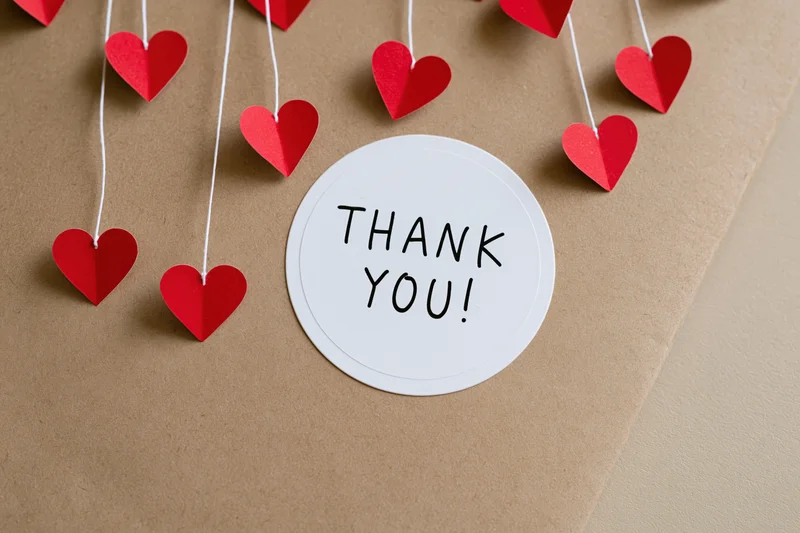 Round white sticker with thank you text on brown paper surrounded by red paper hearts on strings