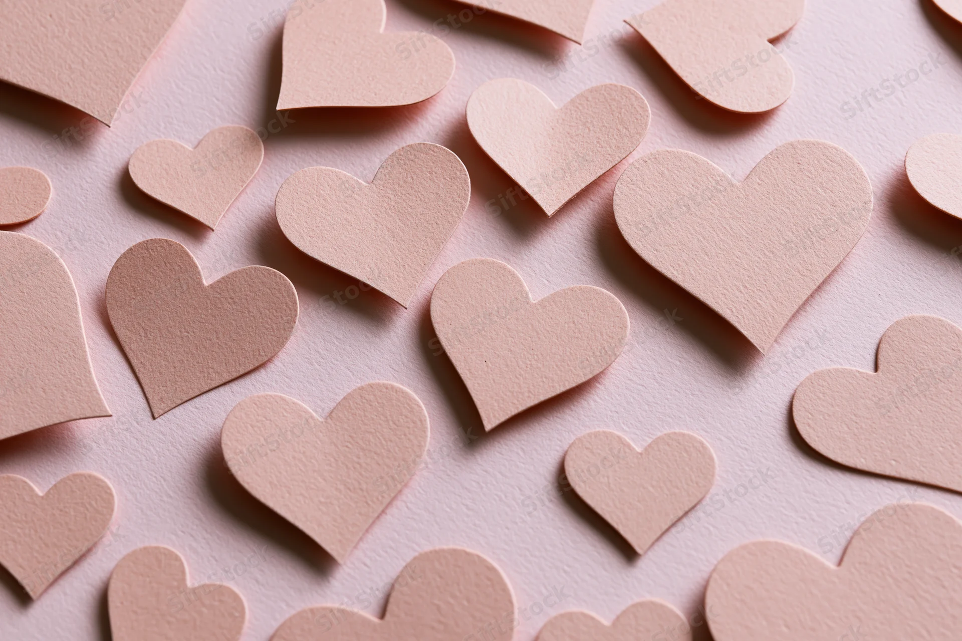 Paper heart cutouts scattered in various sizes on a pink textured surface in flat lay overhead view