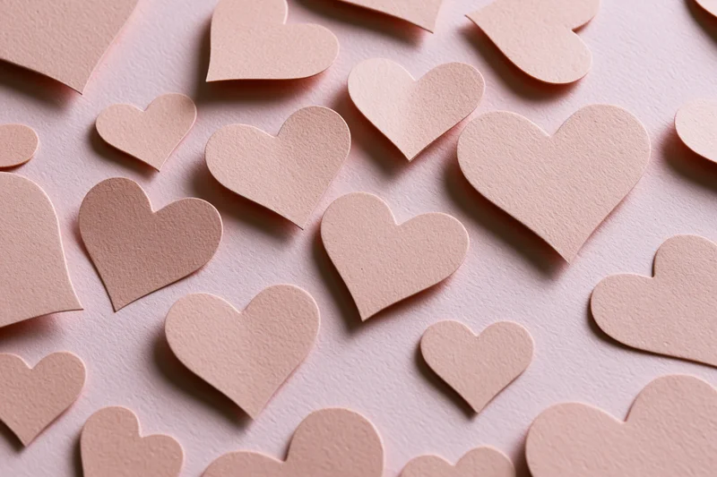 Paper heart cutouts scattered in various sizes on a pink textured surface in flat lay overhead view