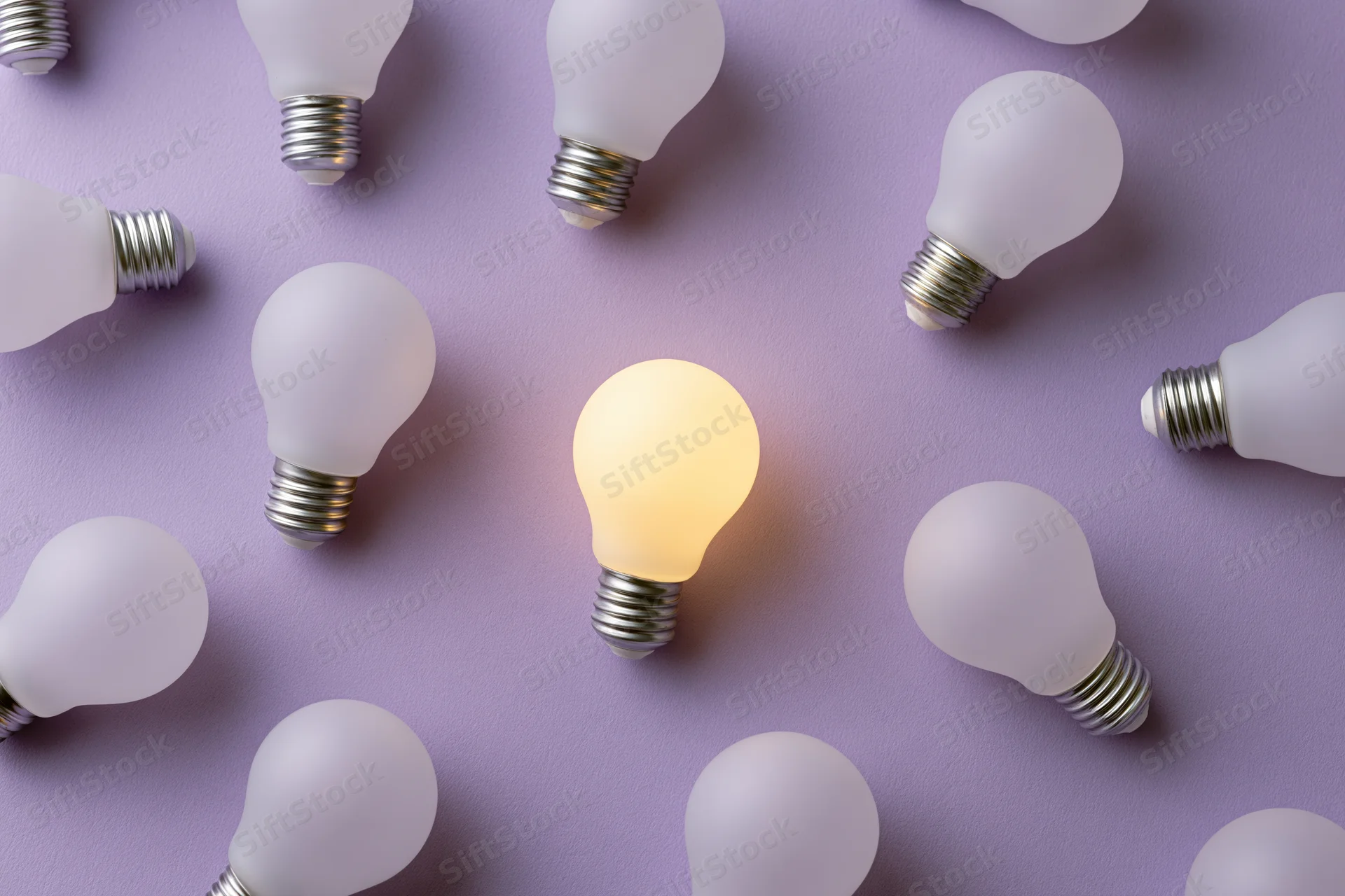 Multiple white light bulbs scattered on purple surface with one glowing in center, flat lay overhead view