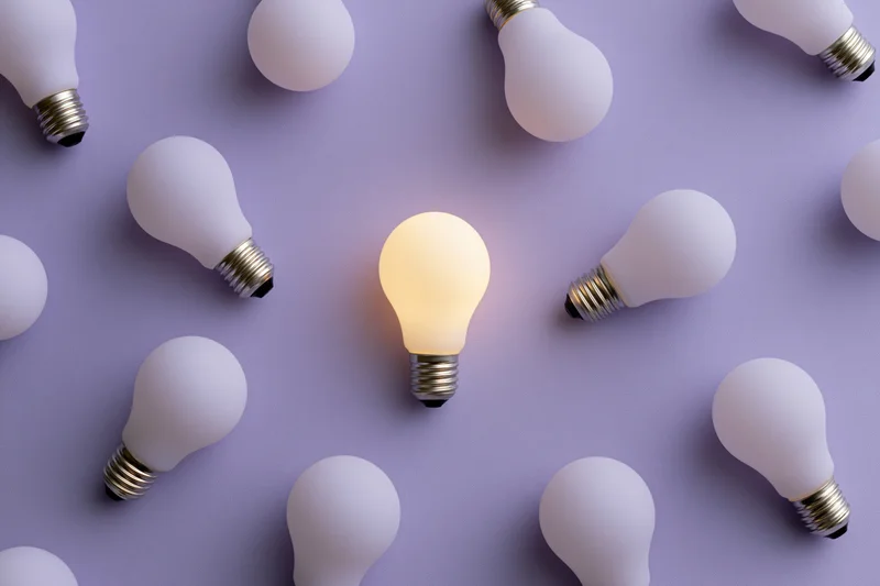 Single glowing lightbulb among unlit bulbs on purple surface