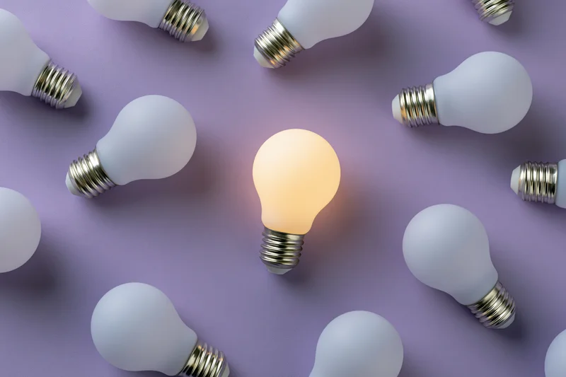 One glowing lightbulb among unlit bulbs on purple surface in flat lay arrangement
