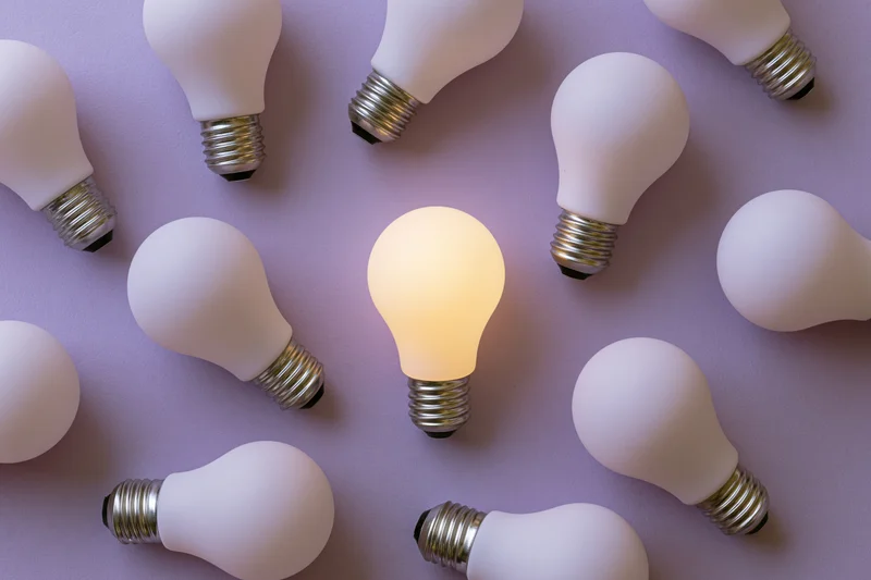 Multiple white light bulbs scattered on purple surface with one illuminated in center