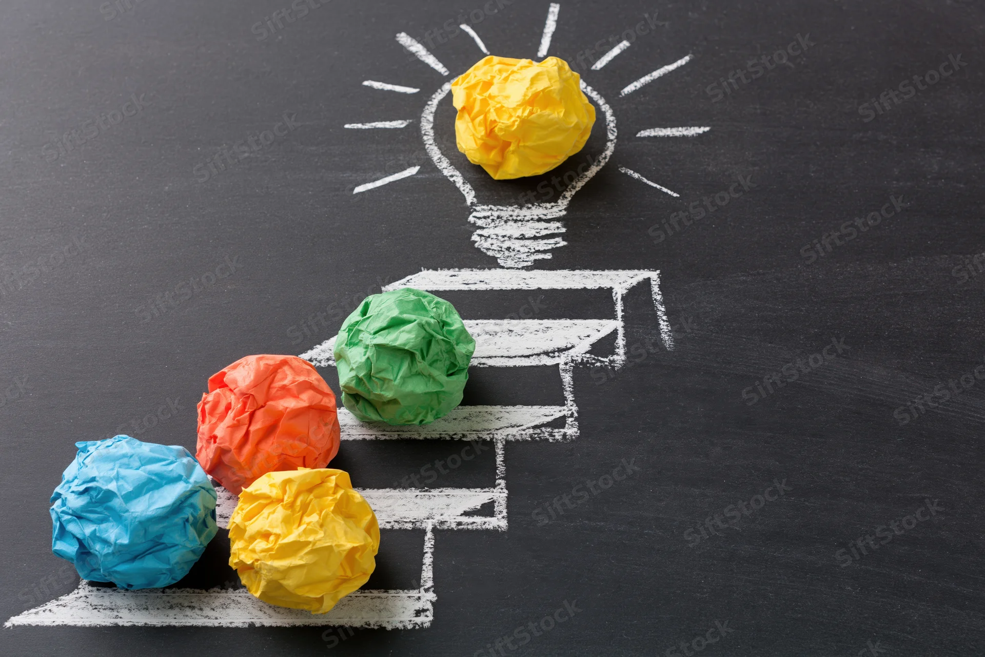 Crumpled paper balls arranged on chalk-drawn steps leading to a lightbulb on a blackboard