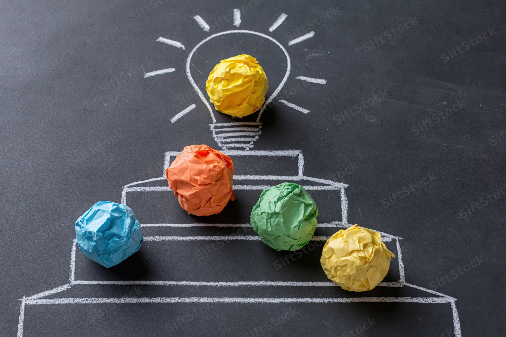 Crumpled paper balls arranged on chalk-drawn steps leading to a lightbulb on a blackboard