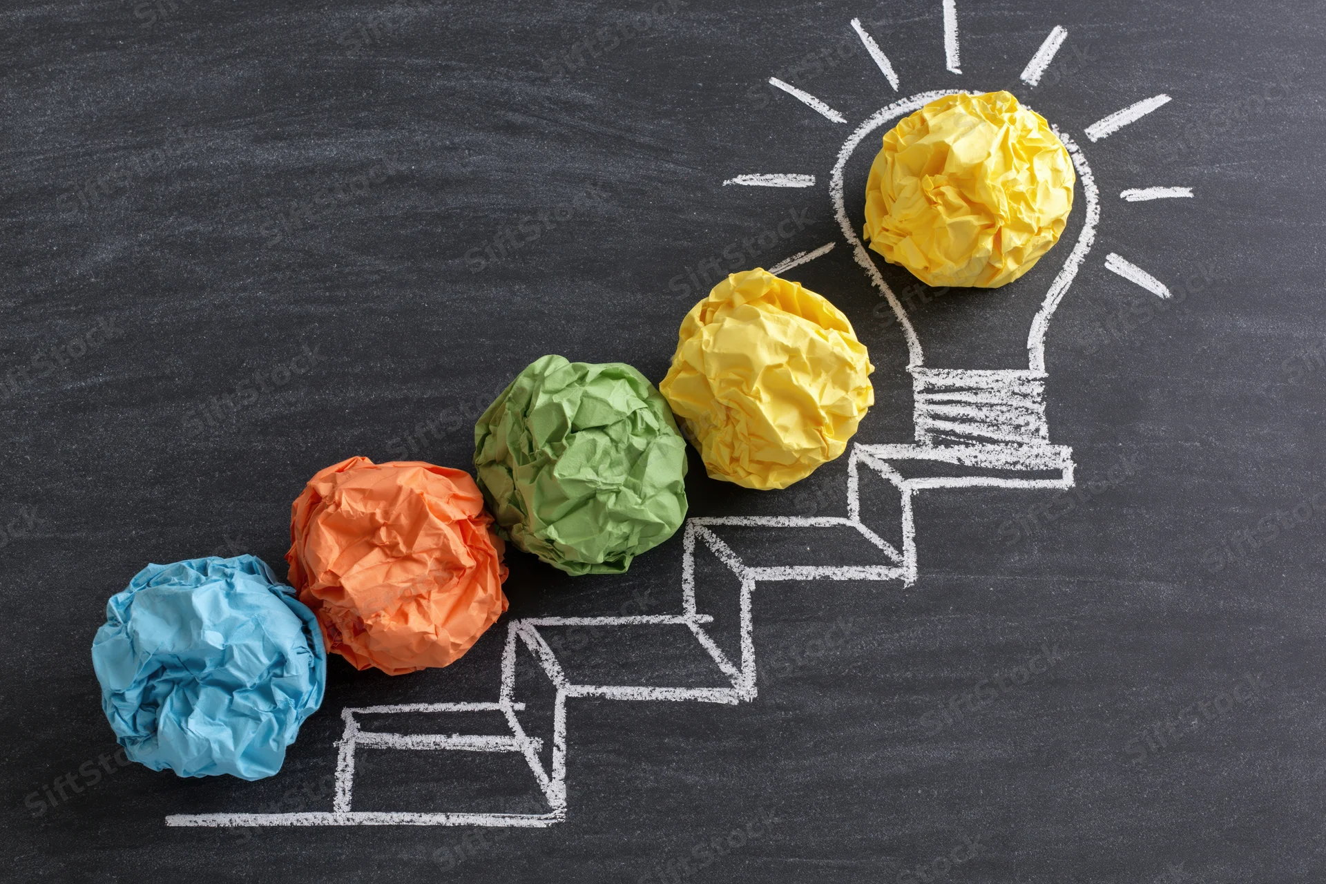 Crumpled paper balls arranged on chalk-drawn steps leading to a lightbulb on a blackboard