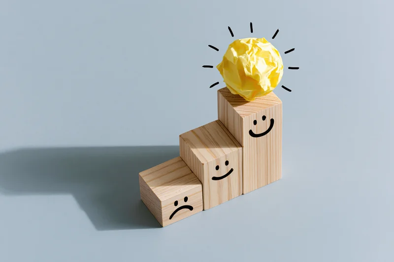 Wooden blocks arranged in ascending staircase with smiley faces and a crumpled yellow paper ball as a lightbulb on top