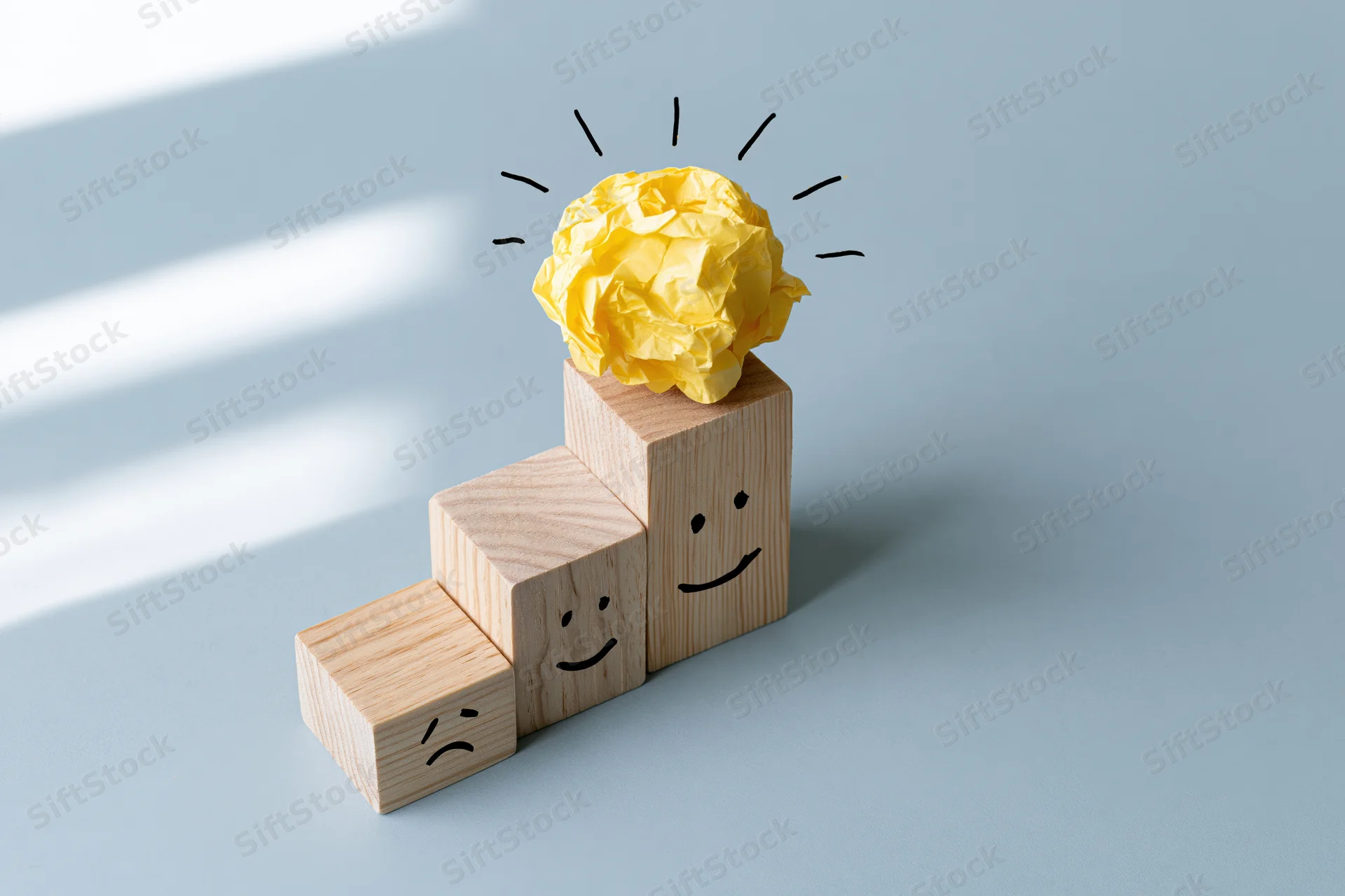 Wooden blocks arranged in ascending staircase with drawn faces and a crumpled yellow paper lightbulb on top