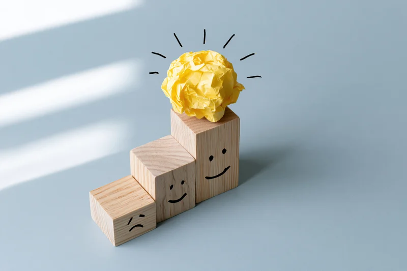 Wooden blocks arranged in ascending staircase with drawn faces and a crumpled yellow paper lightbulb on top