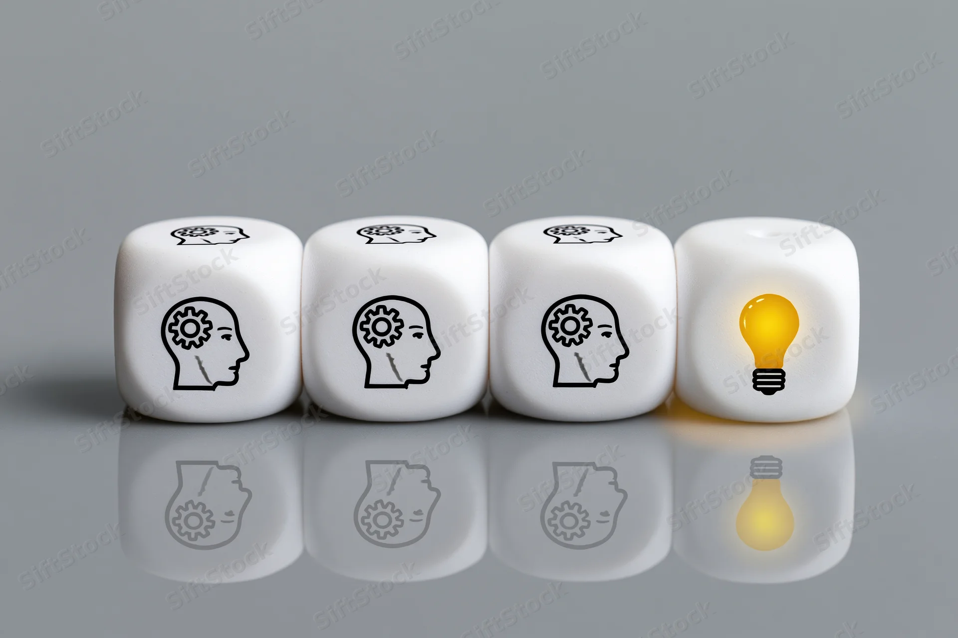 Four white dice in a row with brain symbols and one glowing lightbulb on a reflective gray surface