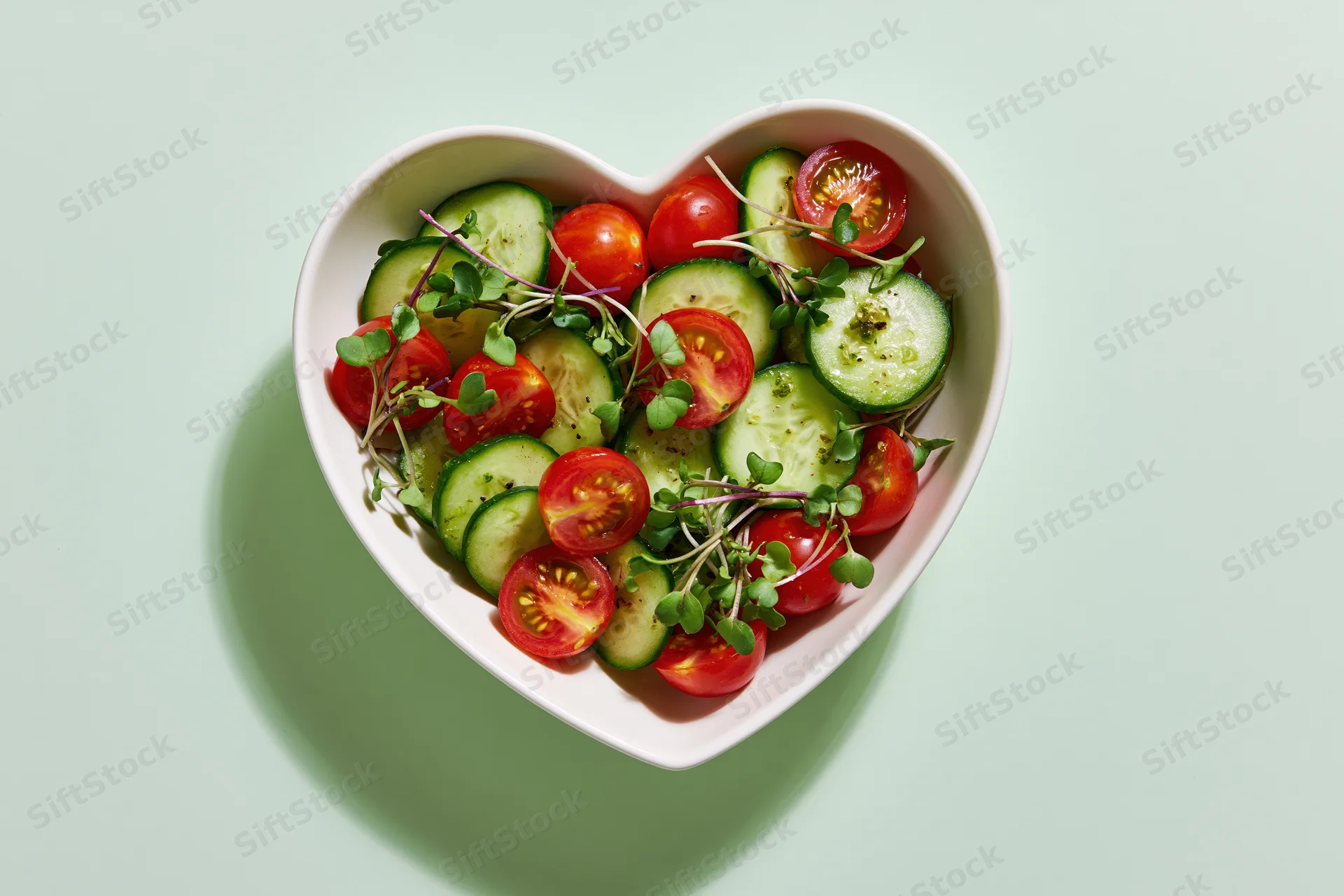 Heart-shaped bowl filled with sliced cucumbers and halved cherry tomatoes on a light green surface