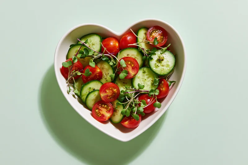 Heart-shaped bowl filled with sliced cucumbers and halved cherry tomatoes on a light green surface
