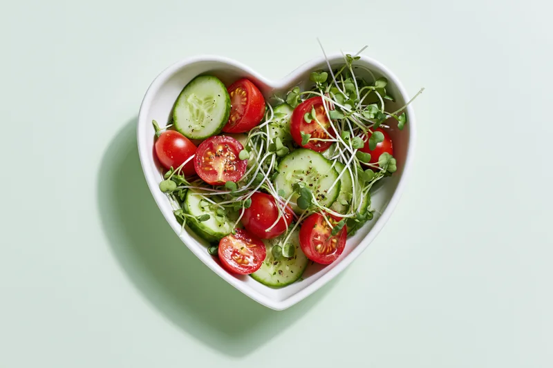 Heart-shaped bowl filled with sliced cucumbers, halved cherry tomatoes, and microgreens on a light green surface