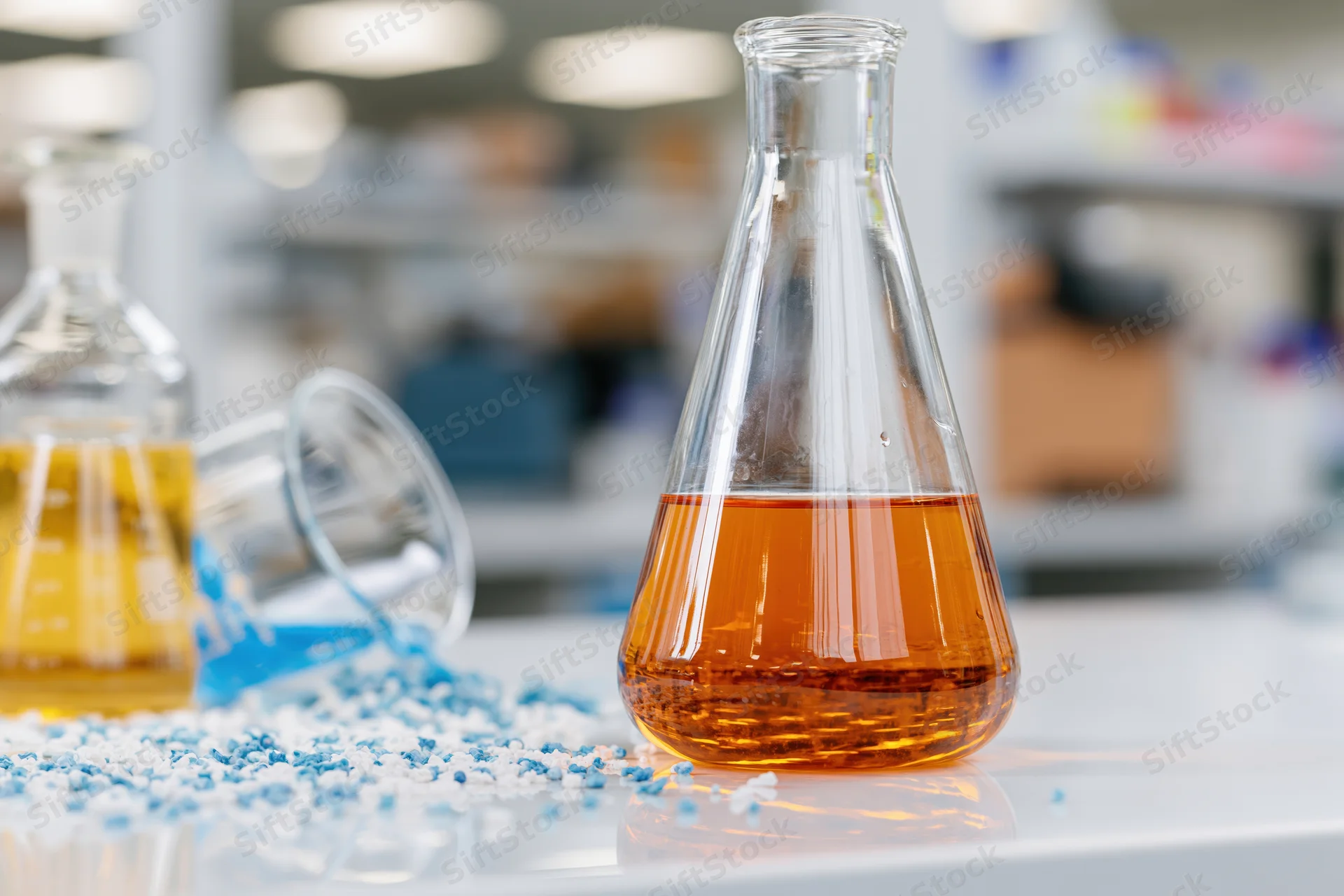Conical glass flask filled with amber liquid on lab bench with scattered blue crystals and another beaker in background