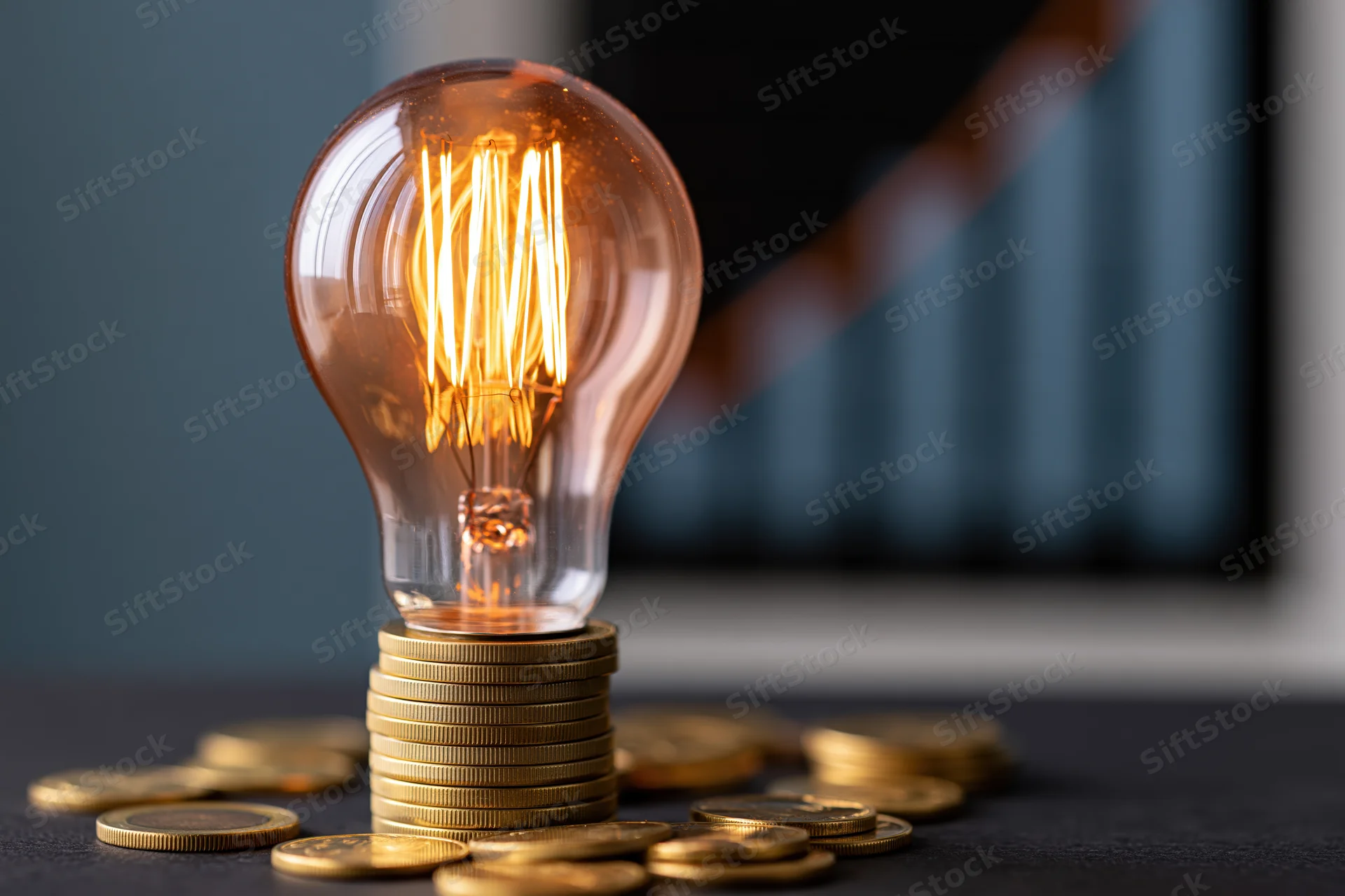 Glowing incandescent lightbulb resting on a stack of gold coins on a dark surface