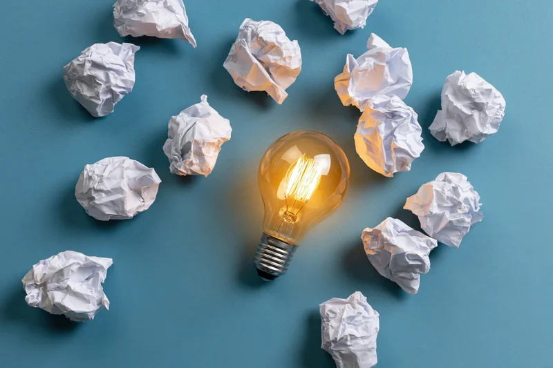 Glowing lightbulb surrounded by crumpled paper on blue surface