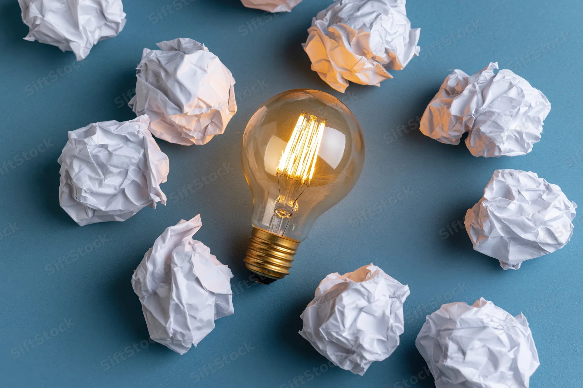 Glowing incandescent light bulb surrounded by crumpled white paper on blue background