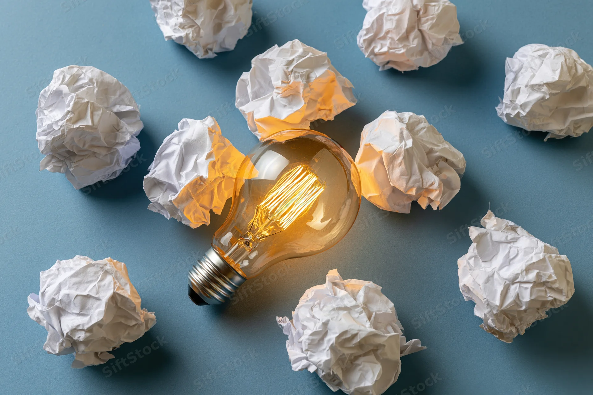 Glowing incandescent lightbulb surrounded by crumpled white paper on blue surface