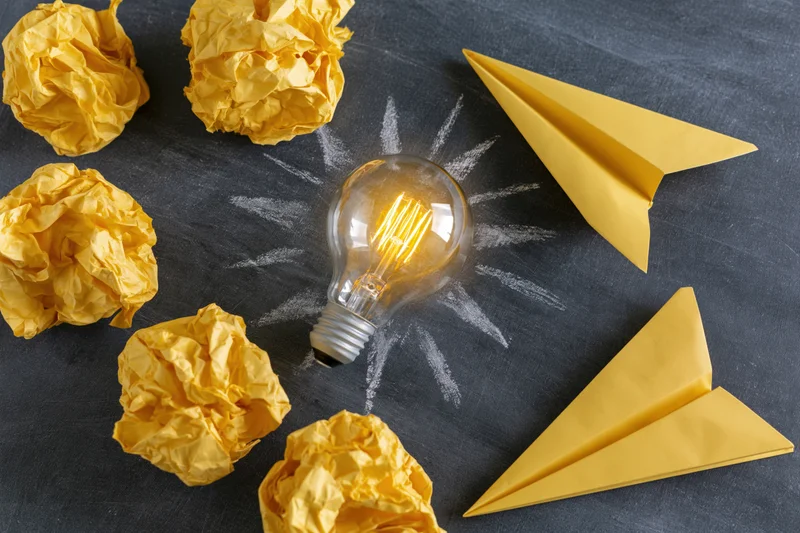 Glowing lightbulb with crumpled yellow paper and paper airplanes on a chalkboard surface