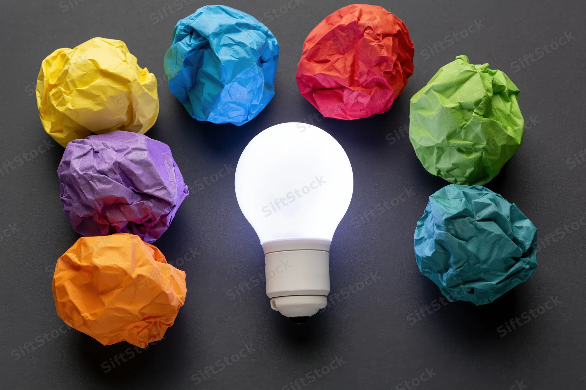 Glowing white led lightbulb surrounded by colorful crumpled paper balls on dark surface