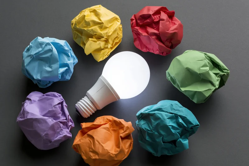 Glowing led lightbulb surrounded by colorful crumpled paper balls on dark surface