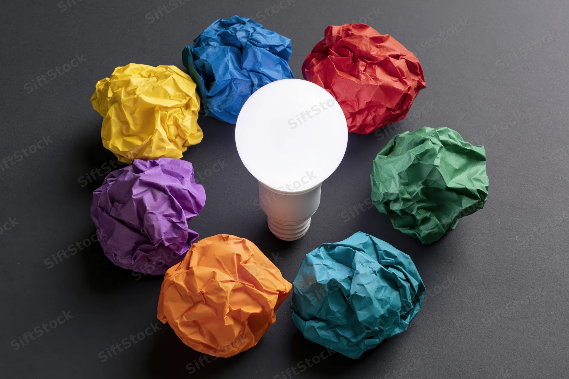 White led lightbulb surrounded by six crumpled colorful paper balls on dark surface