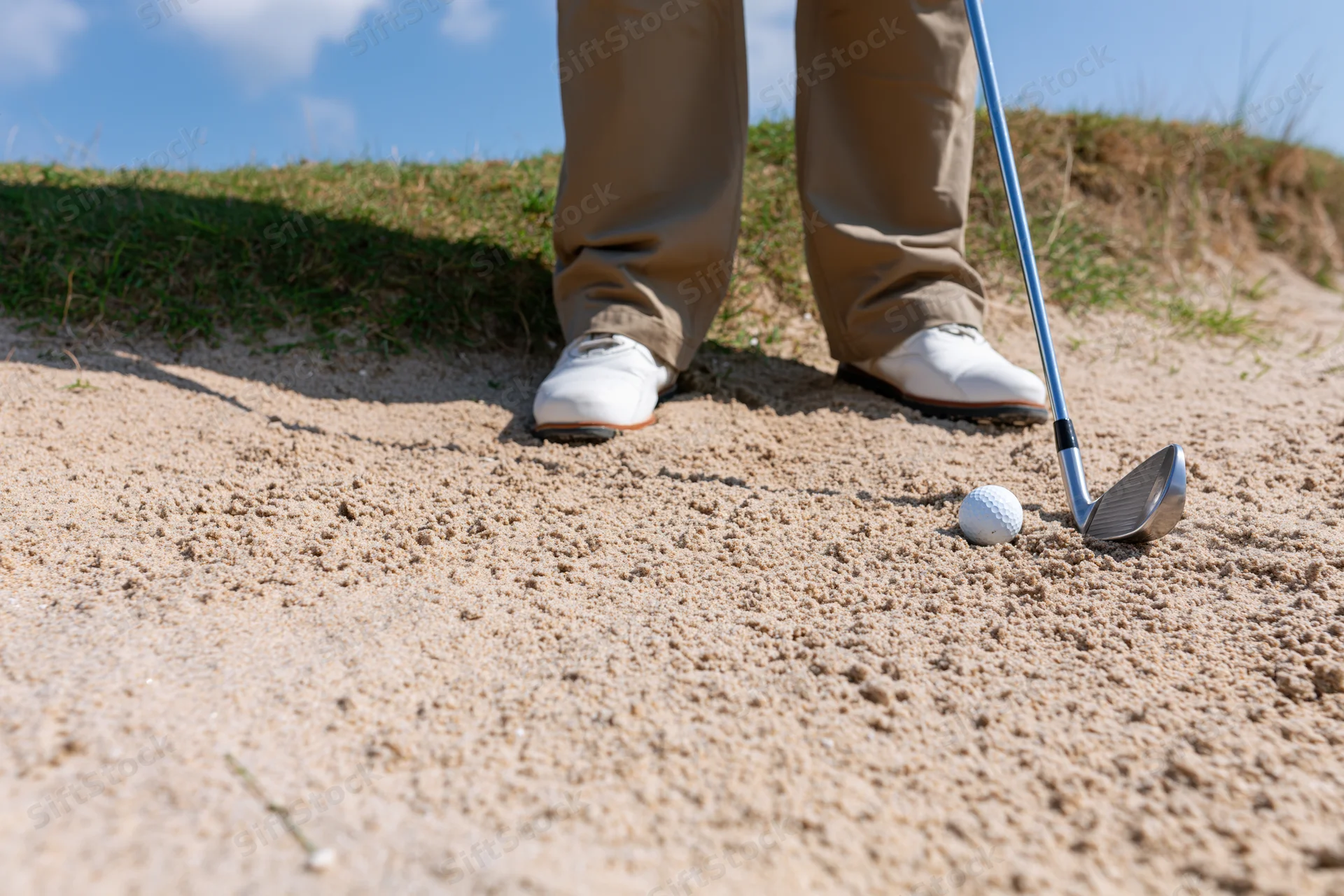 Golfer standing in sand bunker with iron club and golf ball beside feet on course