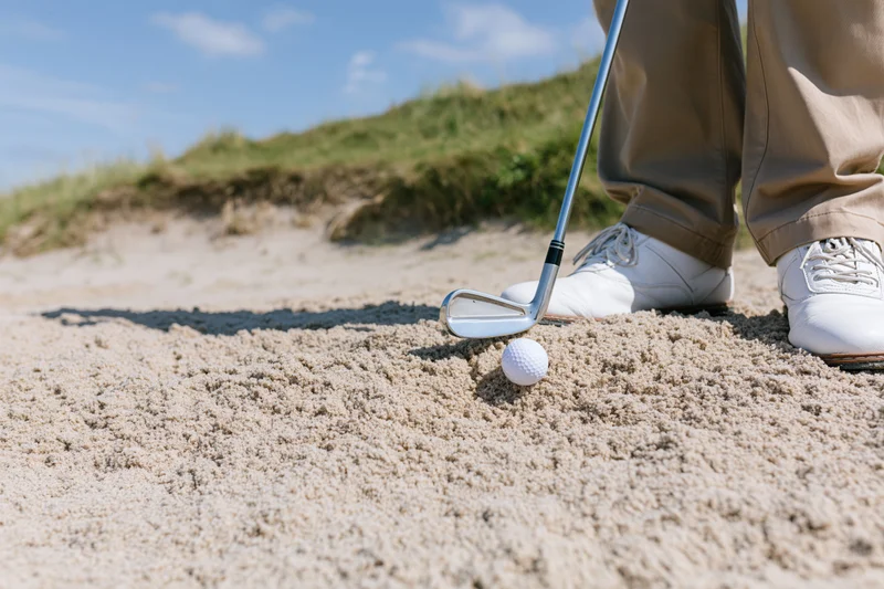 Golfer standing in sand bunker with iron club head near white golf ball on course