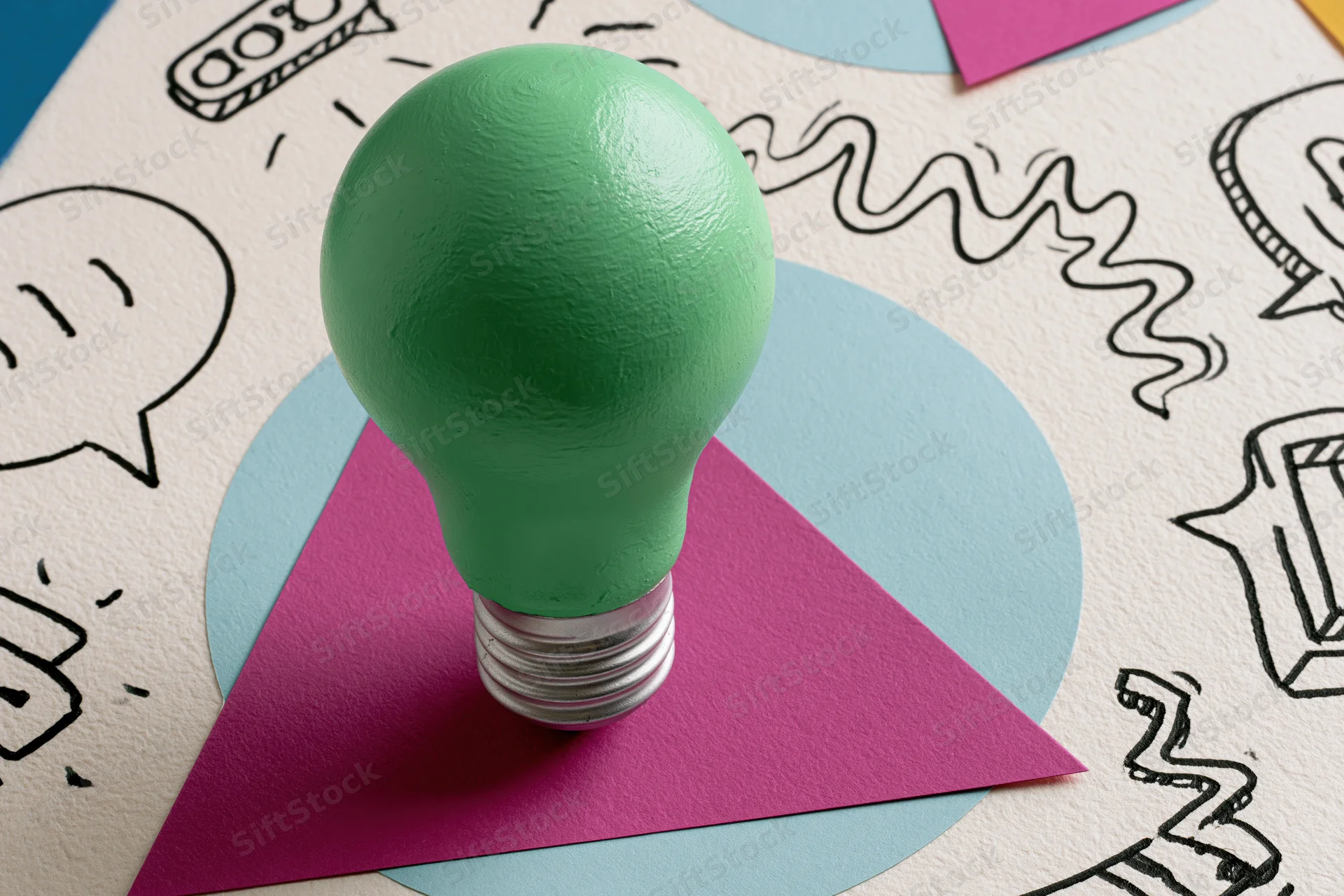 Green painted lightbulb standing on colorful paper shapes with hand-drawn sketches around it