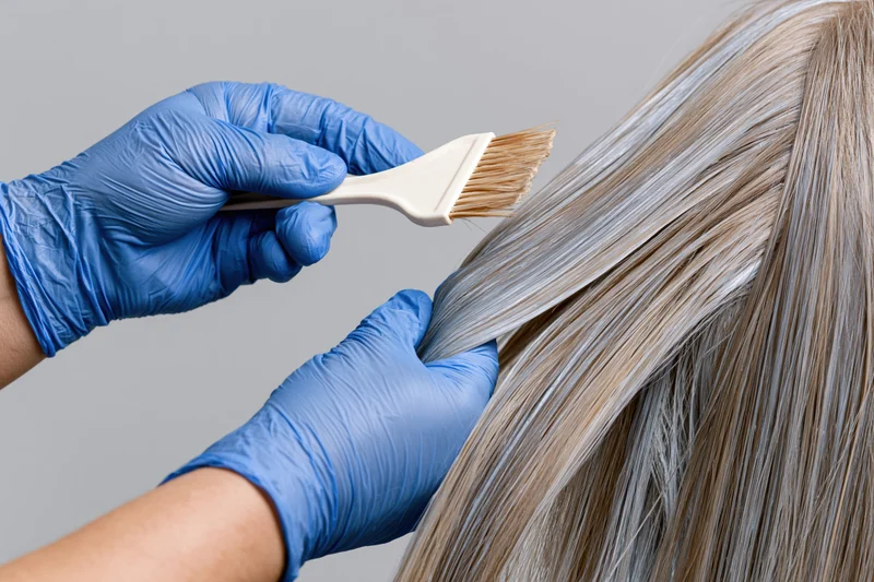 Gloved hands applying hair dye to a section of long blonde hair