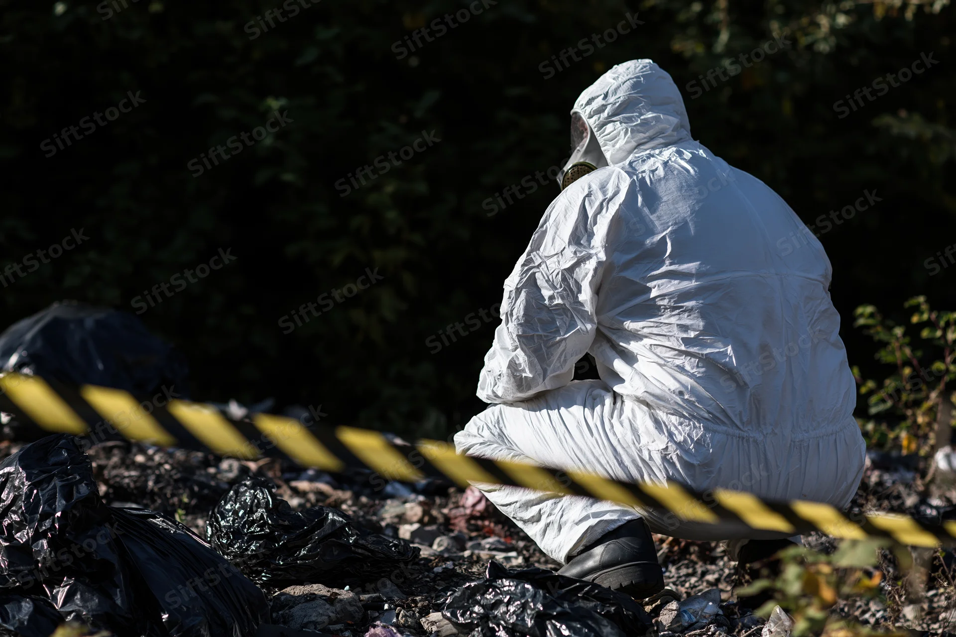Hazmat worker in white protective suit crouching at contaminated outdoor site