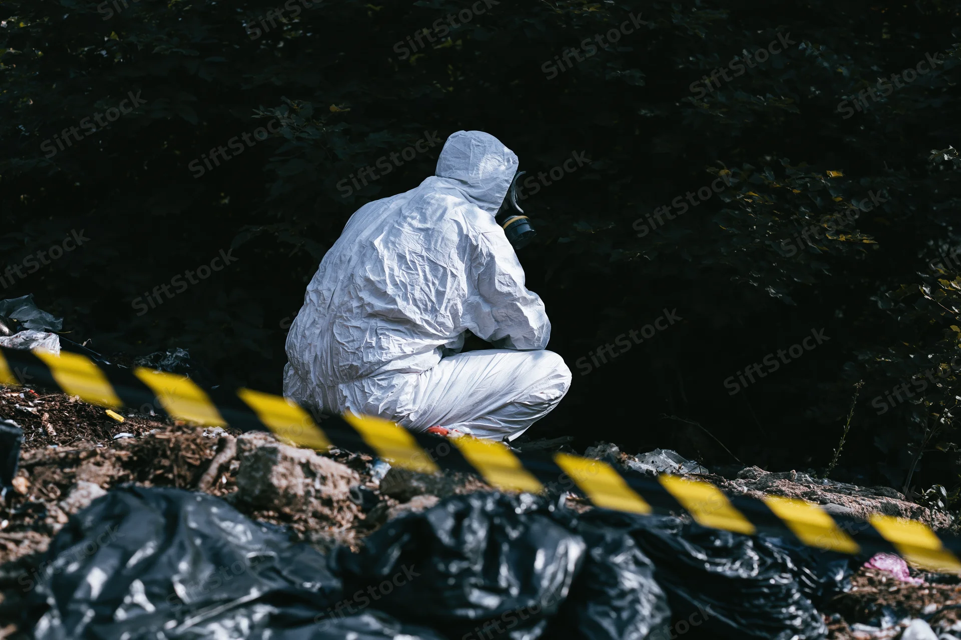 Hazmat worker in white protective suit and gas mask crouching at contaminated outdoor site with caution tape and waste bags