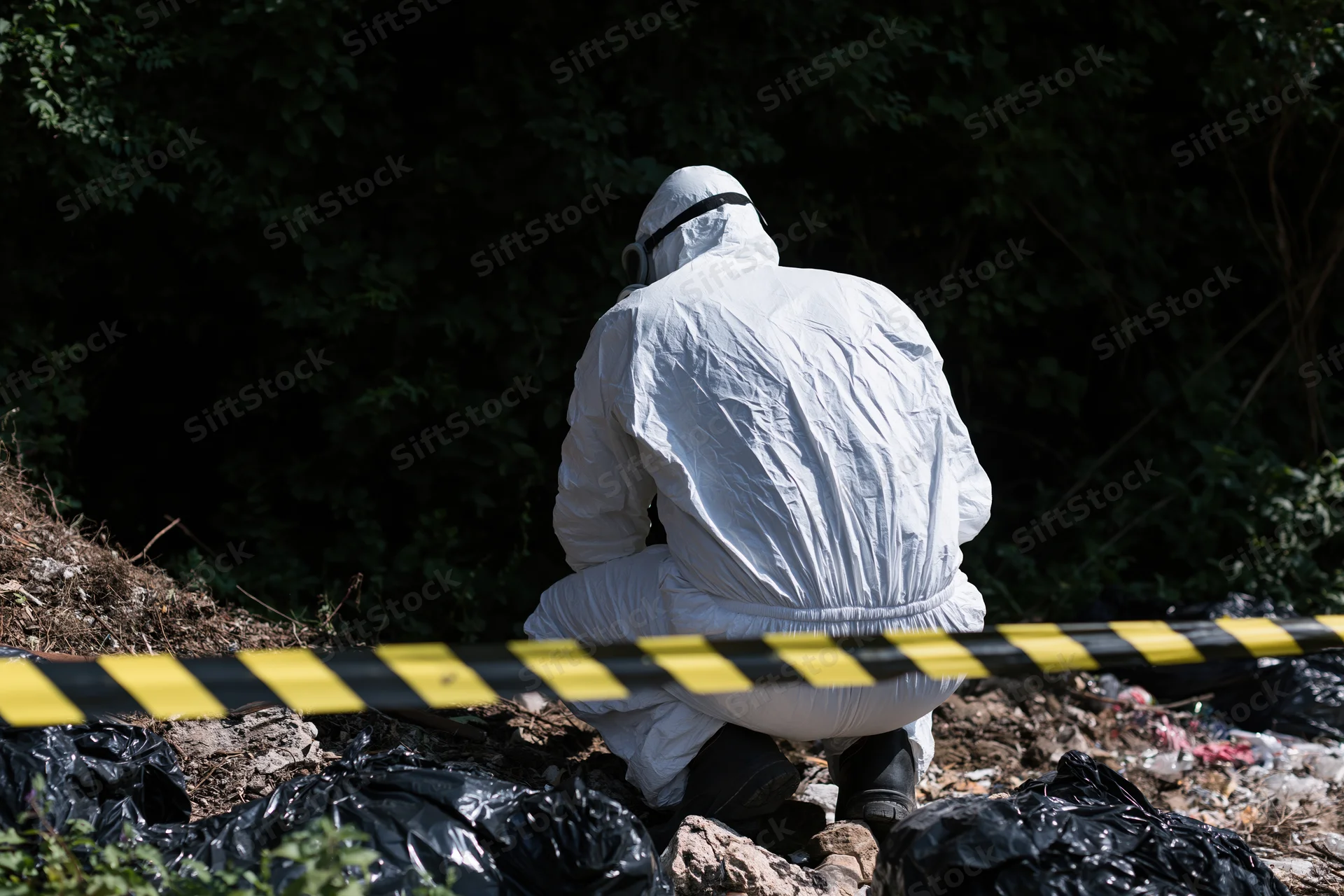 Hazmat worker in white protective suit crouching at contaminated outdoor site