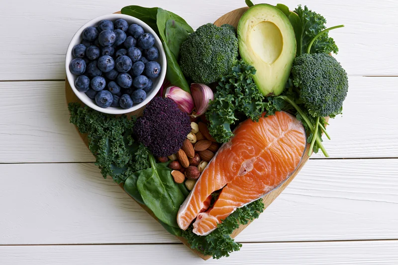 Heart-shaped platter of salmon, broccoli, kale, blueberries, avocado, nuts, and garlic on white wooden surface