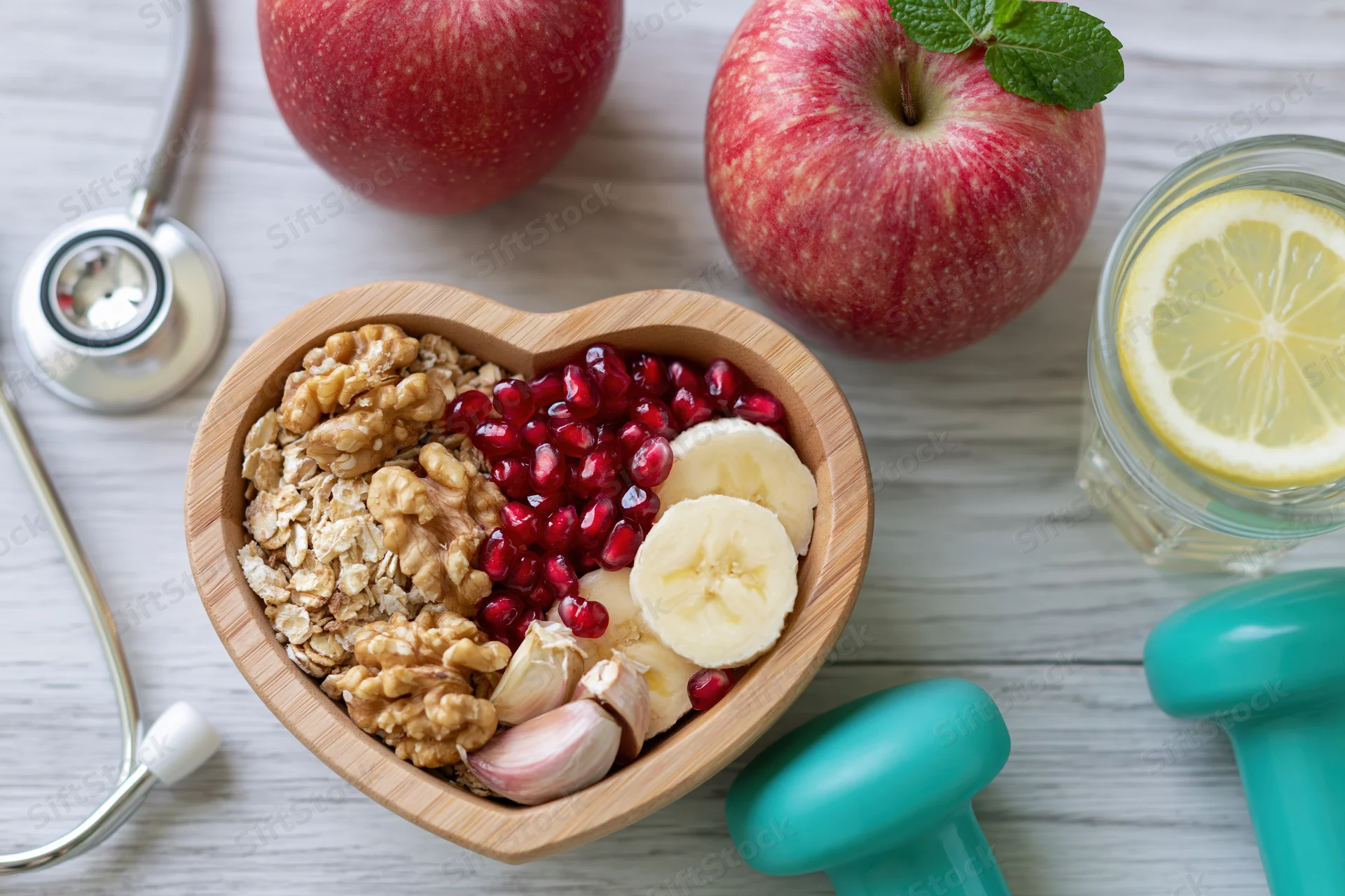 Heart-shaped wooden bowl filled with oats, walnuts, pomegranate, banana, and garlic on a white table with a stethoscope and dumbbells nearby
