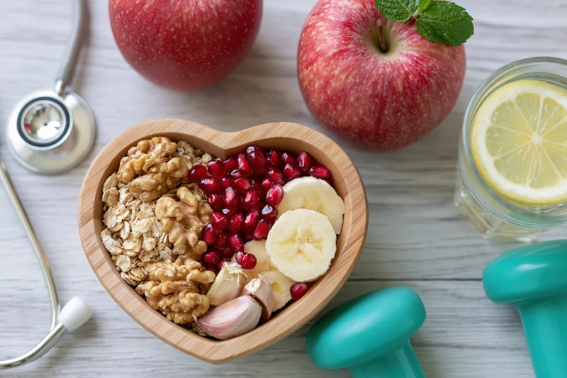 Heart-shaped wooden bowl filled with oats, walnuts, pomegranate, banana, and garlic on a white table with a stethoscope and dumbbells nearby